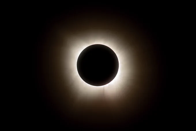 A total solar eclipse with the Moon completely covering the Sun, leaving a bright, white corona radiating outward against a dark sky. The circular silhouette of the Moon is sharply defined at the center, with delicate coronal rays extending in all directions. Photo Credit: (NASA/Jordan Salkin)