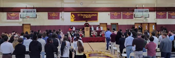group of students and families sitting in gym at Honors Convocation with President Daly speaking onstage
