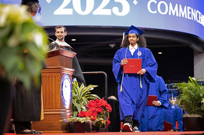 A male Duquesne graduate wearing a blue cap and gown and holding their red diploma walks across the stage at Commencement. 