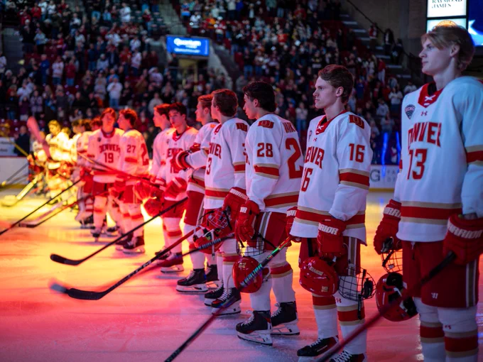 A line of hockey players in red and white uniforms stand on the ice, holding their sticks. The jerseys display the name "Denver." A crowd fills the arena in the background, and dynamic lighting casts a warm glow on the scene.