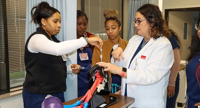 A group of people in a medical setting are gathered around a table observing a demonstration with anatomical models. One person, wearing a white coat, is leading the demonstration. There are various medical props on the table, and the group is attentively focusing on the activity. The setting suggests a learning or training environment.