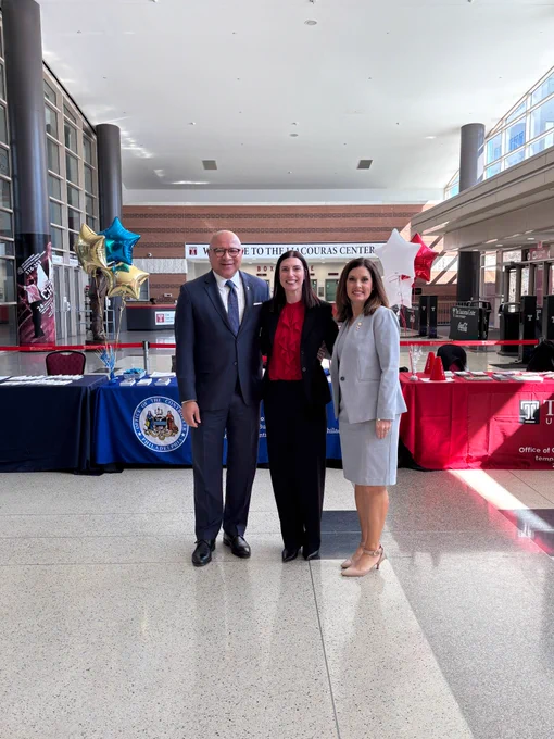 A man in a suit standing with two women, tables with banners on it, balloon in background
