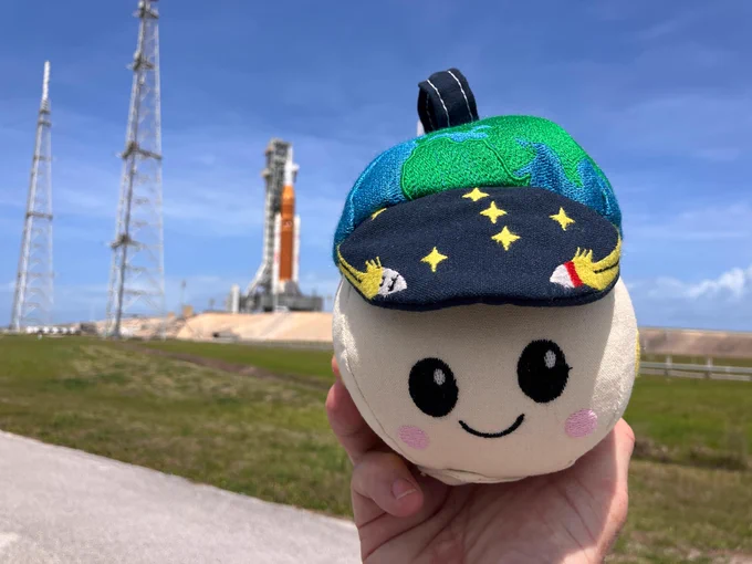 The Rise plushie \u2014 the zero gravity indicator for the Artemis II mission \u2014 smiles at the camera while being held up by someone's hand. In the background, the Artemis II rocket and spacecraft can be seen on Launch Pad 39B at NASA's Kennedy Space Center. Credit: NASA
