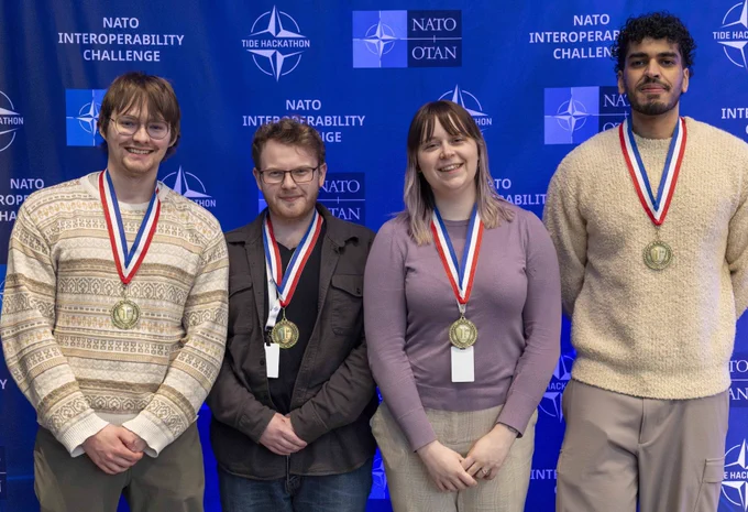 Four individuals wearing medals stand in front of a NATO Interoperability Challenge backdrop at the TIDE Hackathon event. 