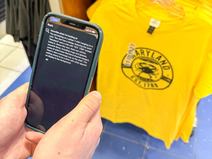 An individual holds a phone utilizing an assistive app with text describing a t-shirt on a rack inside a store at BWI Thurgood Marshall Airport.