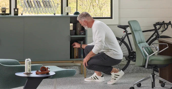 Man in casual clothes crouching to open a lower cabinet door in a modern office with a bicycle and a Massaud Executive Office Chair nearby.