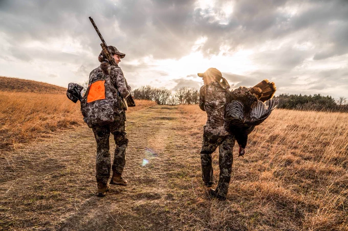 Two individuals dressed in camouflage gear walk on a grassy path in an open field. One carries a turkey over their shoulder, and the other has a shotgun slung across their back. The sun is setting, casting a warm glow over the landscape.