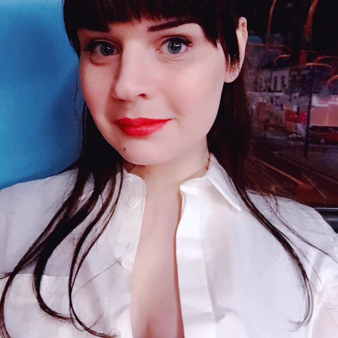 Selfie. A young woman in a crisp white shirt, undone to show a bit of cleavage. Her hair is long, and almost wispy, dark brown with blunt bangs. No glasses, just blue eyes and a bold red lip.