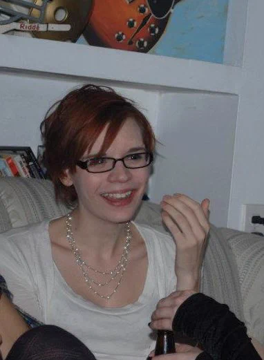 A you g trans woman, too thin, like so many of us are at that age, in a white outfit wearing a complicated beaded necklace, sitting on a couch and laughing. She has short, red hair with swooping bangs. She is laughing. Eyebrows less intense. Someone nearby is holding a beer bottle.
