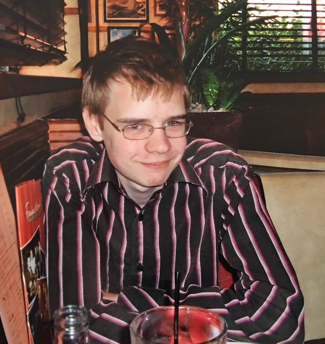 Old, analog photo: What looks like a young man (you have to say that because the person in the picture turned out to be a trans woman) sitting in a booth at a Frankie and Bennys. The person has very short, blonde hair, wire-rimmed glasses, thick eyebrows, and is wearing a black collared shirt with pink pinstripes