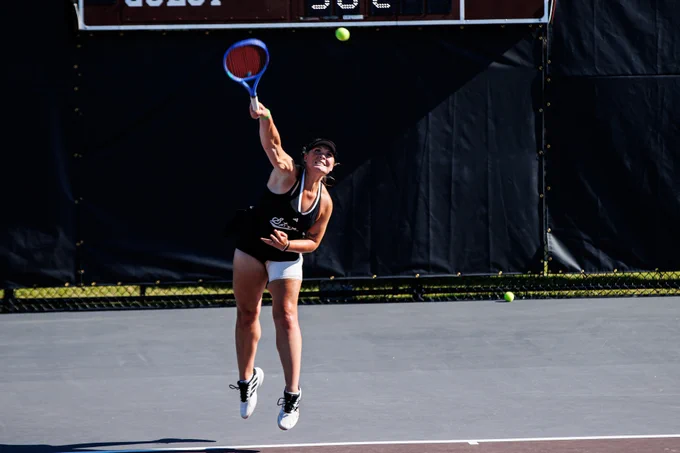 A women’s tennis player leaps into the air to hit an overhead shot, eyes focused on the ball above her racket on an outdoor court.
