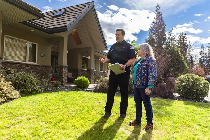 A person in uniform holding a clipboard stands with another person on a grassy lawn in front of a suburban house. The sky is partly cloudy and trees are in the background.