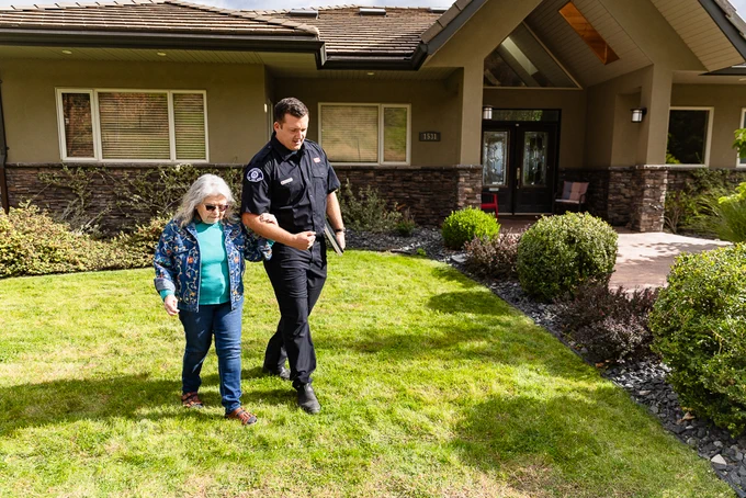 A person in a uniform helps another person walk across a grassy lawn in front of a house. The house has stone accents and tall windows. The scene is well-lit and appears to be taken during the day.