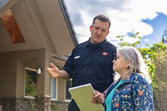 A person in an official uniform with a Canadian flag patch is talking to another person outside a house. The person in uniform is holding a large notepad or document.