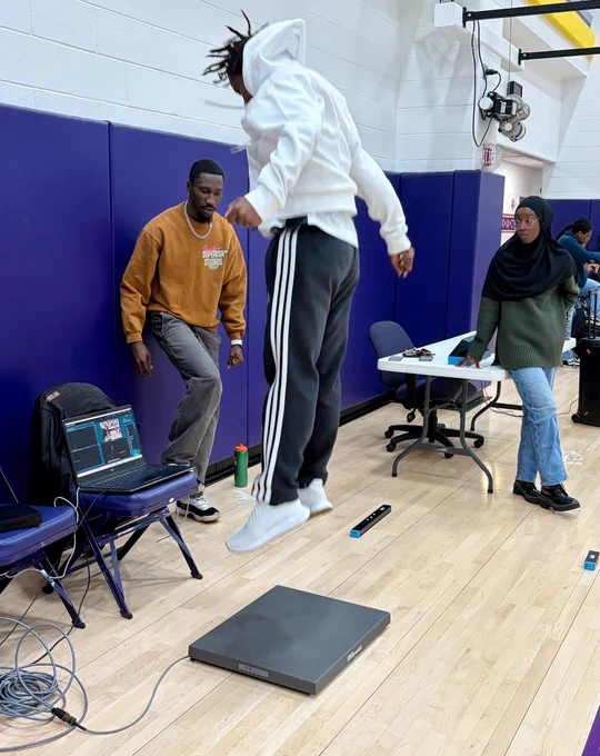 A person is jumping on a performance measuring platform in a gym. Two other individuals stand nearby, observing. A laptop and other equipment are visible on a table in the background.