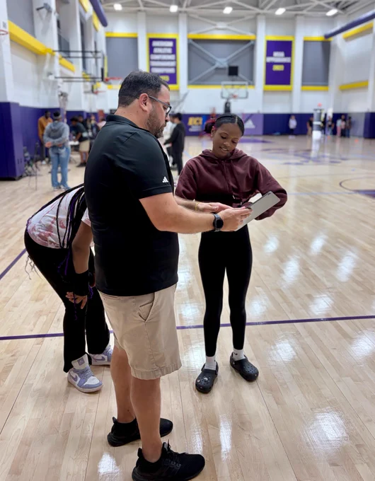 In a gymnasium, two people are having a conversation while holding a tablet. The gym has a hardwood floor and basketball hoops.