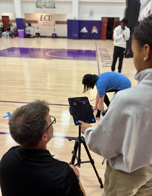 A person kneels on a basketball court holding a device on a tripod, while another person bends over in the background adjusting something on the floor. The court has purple accents.