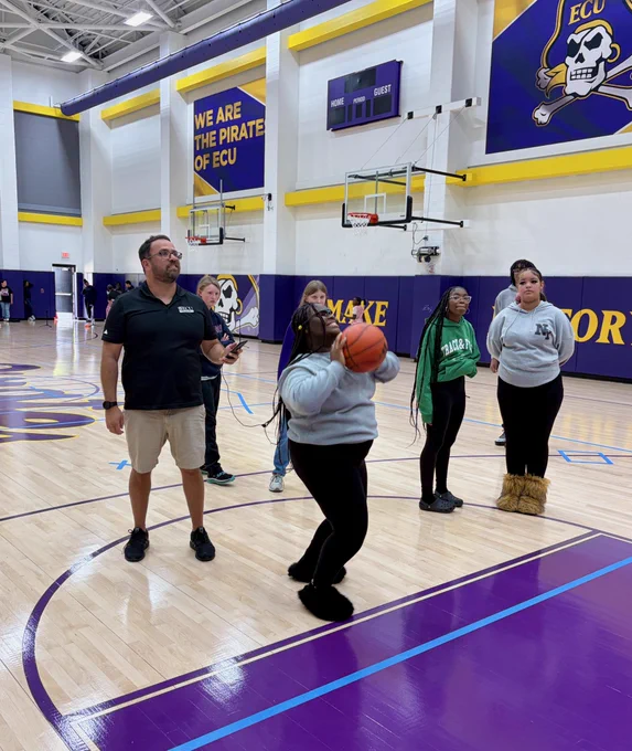 A group of people are in a basketball court. One person is shooting a basketball, while wearing glasses that track eye movement.