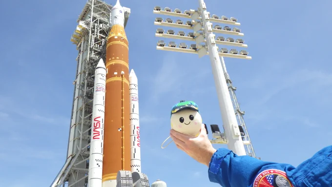 Canadian Space Agency astronaut Jeremy Hansen holds up the \"Rise\" zero gravity indicator plushie in front of the Space Launch System rocket and Orion spacecraft on Launch Pad 39B at NASA's Kennedy Space Center. The orange core stage of the rocket stands between two white solid rocket boosters and is topped with the white Orion spacecraft. A pole with light fixtures stands between Hansen and the rocket. Credit: NASA