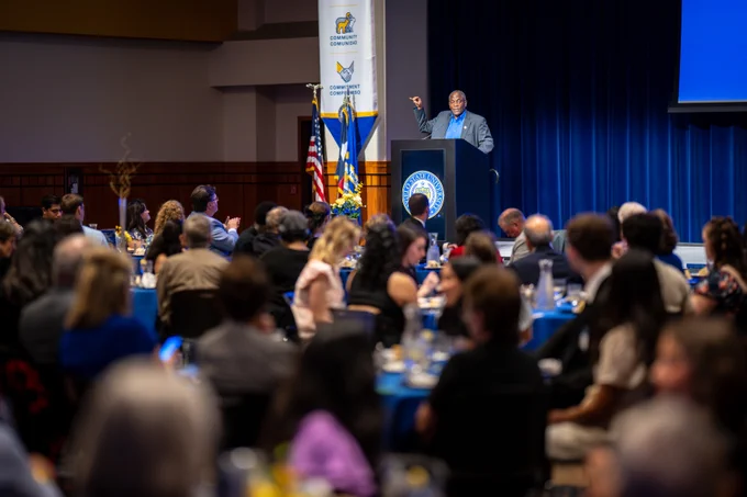 A speaker stands at a podium addressing a room full of seated attendees. The setting is formal, with a blue curtain backdrop and a banner displaying the name "Fresno State University." Tables are arranged with blue tablecloths and dining settings, and a flag is visible near the podium. 