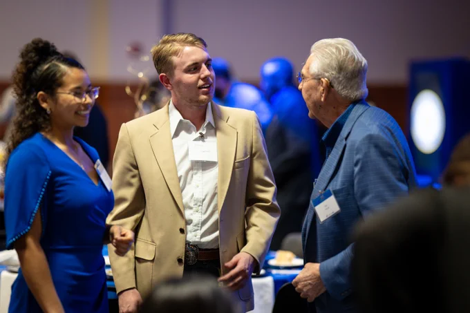 Three people are engaged in conversation at an event. The woman on the left is wearing a blue dress and glasses, smiling. The man in the center has short hair, wears a tan blazer, and has a name tag. The elderly man on the right is dressed in a blue jacket, speaking animatedly.