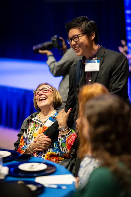 An woman laughs joyfully while sitting at a table. She is engaging with a young man in a suit, smiling at her, creating a warm and lively atmosphere. In the background, a photographer is capturing moments.