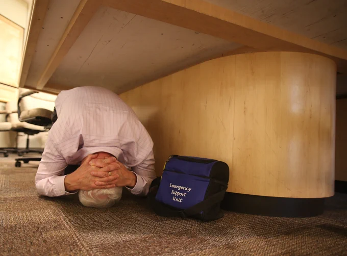 A person under a table holding on to his head with an emergency kit next to him. Drop, cover and hold on is a safety action you can take during earthquakes. 