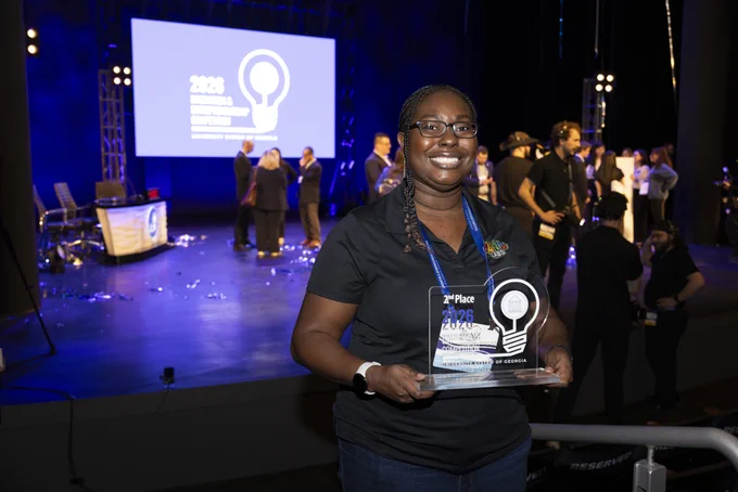 An individual smiles while holding a plaque for winning second place in a competition. In the background, people are gathered on a stage with a large screen.