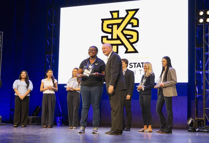 A group of people are on stage at an event. One individual in the center, holding an award, stands next to another who is smiling. Behind them, a large screen displays the Kennesaw State University logo. More people are standing in line.