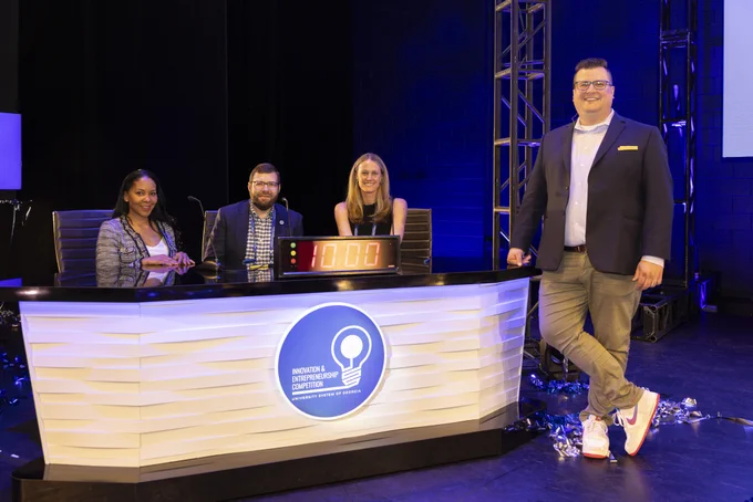 A group of four individuals is standing and sitting around a large desk on a stage. The desk has a sign that says "Innovators" atop it. The setting is well-lit, with metal scaffolding and black curtains in the background. The people are smiling, appearing to be part of an event or competition.