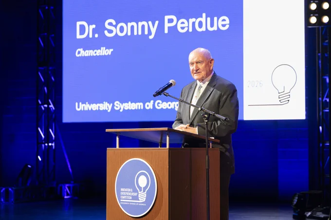 Sonny Perdue stands at a podium on a stage with a microphone. Behind, a screen shows the text "Dr. Sonny Perdue, Chancellor, University System of Georgia." The stage is lit with blue and white lights.