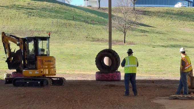 Two workers in yellow vests and white hats stand on a dirt field near a yellow excavator. A large tire sits on a red platform.
