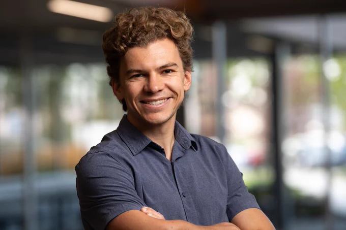 A smiling person with curly hair, wearing a short-sleeve button-up shirt, stands with his arms crossed in a softly lit indoor setting.