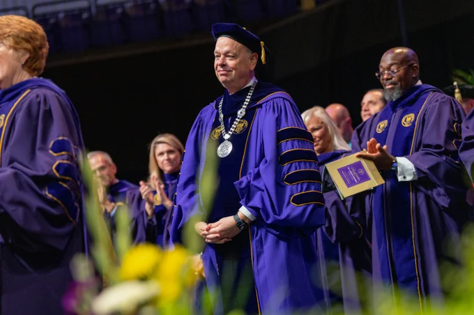 President Jim in his presidential robe wears the silver Presidential Chain of Office.  The chain of office is made of sterling silver and two gems — a golden citrine and a purple amethyst — reflecting JMU’s school colors. The gems are set in the circular sterling silver medallions that are incorporated into the chain. One medallion is textured like bluestone to symbolize the older buildings on JMU’s campus. The other medallion features a brick texture to reflect the university’s newer buildings. The university medallion that hangs from the chain features the seal of James Madison University, the university’s name and the date of its founding, 1908. The design for the JMU seal is based on the coat of arms of the Madison family and contains the words “Knowledge is Liberty,”.