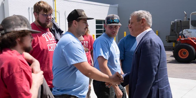 An individual in a suit jacket shakes hands with another individual wearing a baseball cap, jeans and a t-shirt. Four others look on. 