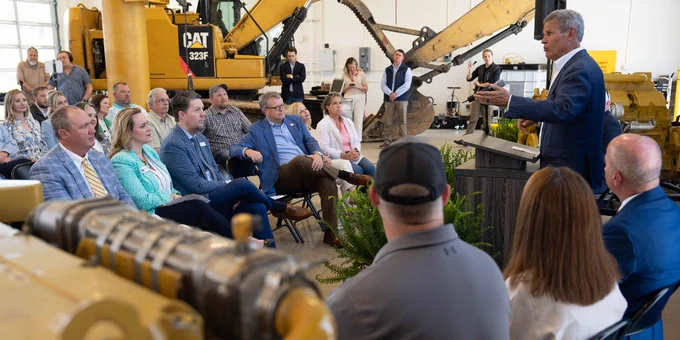 An individual stands behind a podium, addressing a large crowd gathered for a ceremony. Heavy construction equipment is featured in the back.