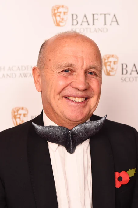 Doug Allan on the red carpet at the BAFTA Scotland Awards. He is smiling at the camera and is wearing a black suit, with a bow tie the shape of a whale tail.