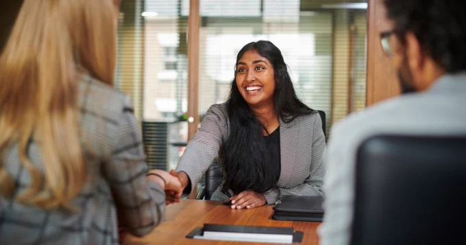 Three professionals in an interview setting, with two greeting each other by smiling and shaking hands
