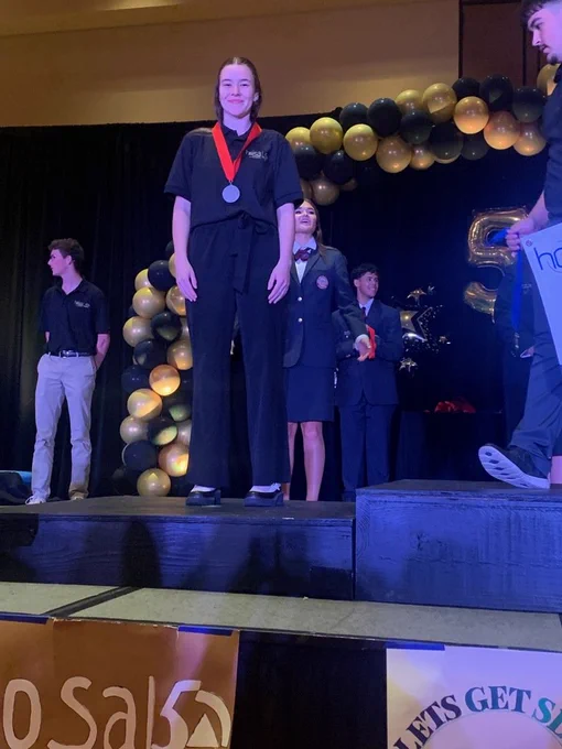 A stage with an individual receiving a medal, standing with others. Behind them, a backdrop of balloons.