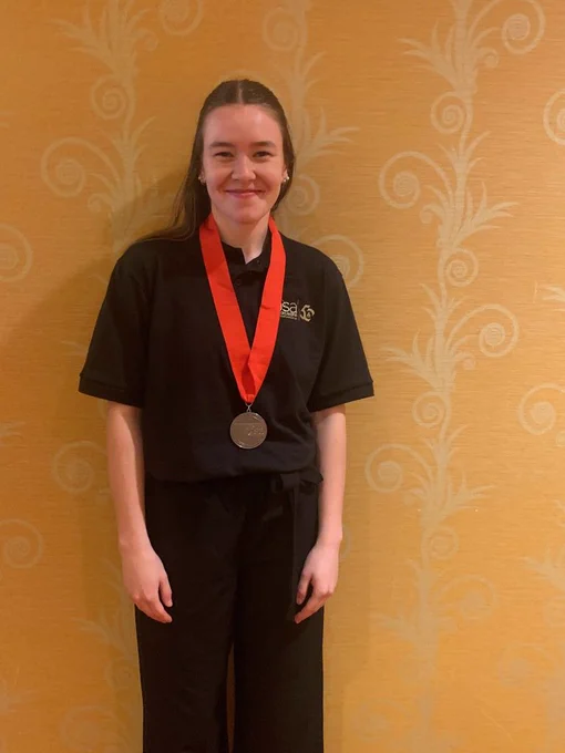 A person in a black shirt and pants stands against a yellow wall with swirly patterns, smiling with a medal around their neck.