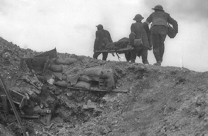 Canadian soldiers carrying a wounded comrade on a stretcher across uneven ground and sandbags on the battlefield at Vimy Ridge.