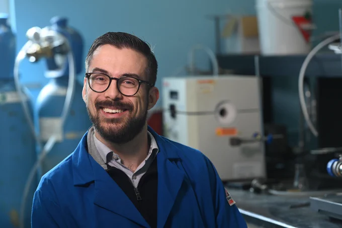 A scientist wearing a blue lab coat with dark hair and beard
