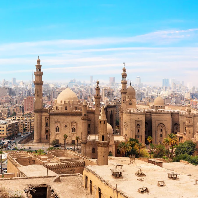  A wide cityscape of Cairo featuring historic Islamic architecture in the foreground—large sandy-colored mosques with domes and tall minarets—set against a dense backdrop of modern buildings under a bright blue sky with light clouds. 