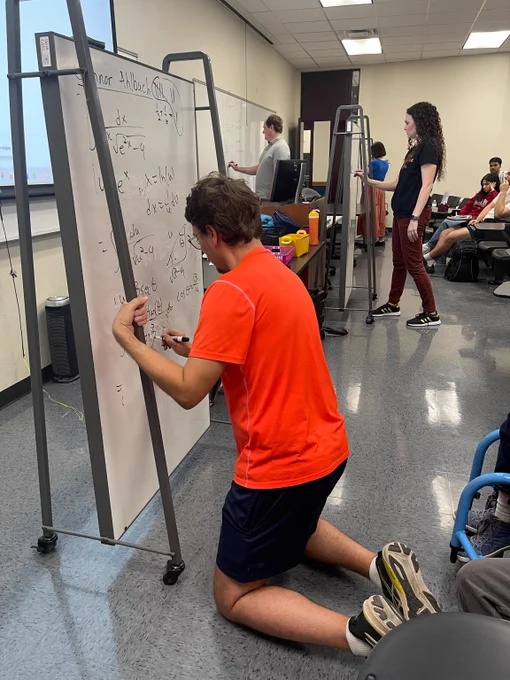 Dr. Ahlbach kneeling and writing complex math equations on a whiteboard in a classroom setting.