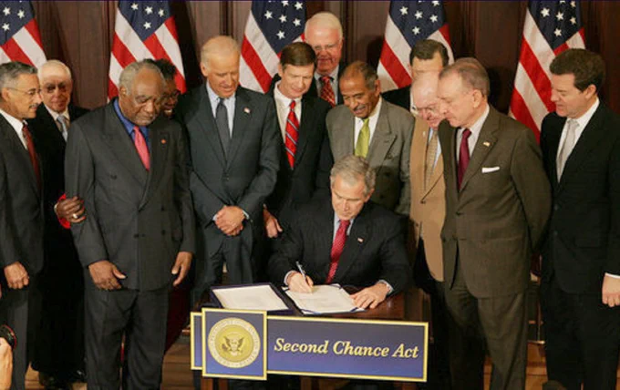 A group of people stand around a table as an individual signs a document labeled "Second Chance Act." Several American flags are displayed in the background.