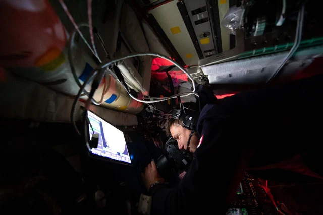 Jeremy Hansen takes a photograph out the window of the Orion spacecraft window. Hansen has the viewfinder of the camera pressed against his face and his left hand is adjusting the lens. Around him, the interior of the spacecraft is dark except for a brightly glowing screen in front of him and some areas of red glow. Credit: NASA