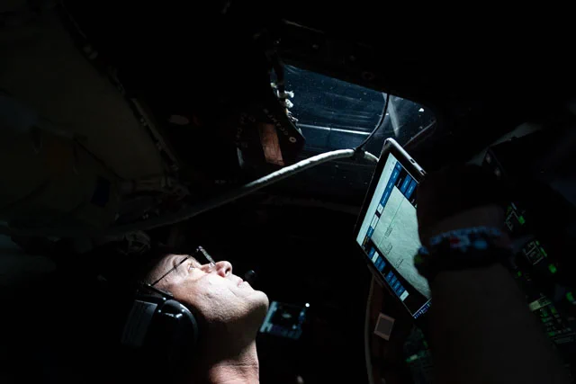 Reid Wiseman looking out a window in the Orion spacecraft. The interior of the spacecraft is mostly dark. Wiseman\u2019s face is brightly lit by a combination of light from outside the window and the glow of a screen in front of him. His face is upturned to look out the window above him. Credit: NASA