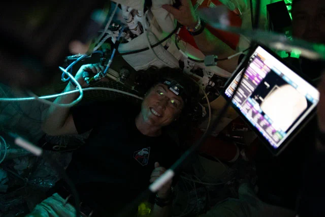 Christina Koch in the Orion spacecraft. She is smiling at the camera and wears glasses and a headlamp. The interior of the spacecraft is mostly dark except for the bright screen of a tablet on the right and some areas that glow blue and green. Credit: NASA
