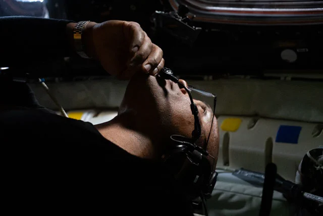 Victor Glover in the Orion spacecraft. His head is tilted back to look out a window above him, obscuring his face from the camera. The interior of the spacecraft is dark. Glover is mostly cast in shadow, except for his side profile. Credit: NASA