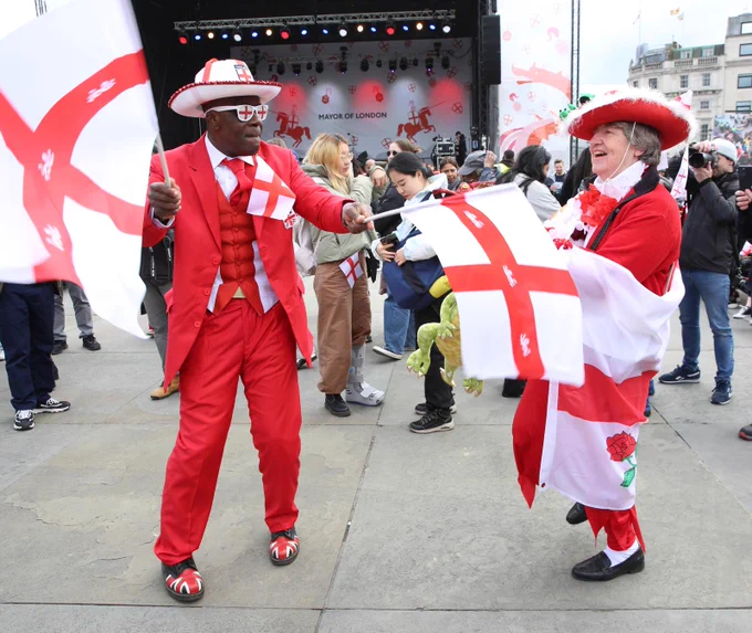 Two people dressed in bright red and white St George’s Day outfits wave large England flags in Trafalgar Square during the St George’s Day Festival. The festival stage is visible in the background, decorated with red illustrations and “Mayor of London” branding. A crowd of attendees is gathered behind them.